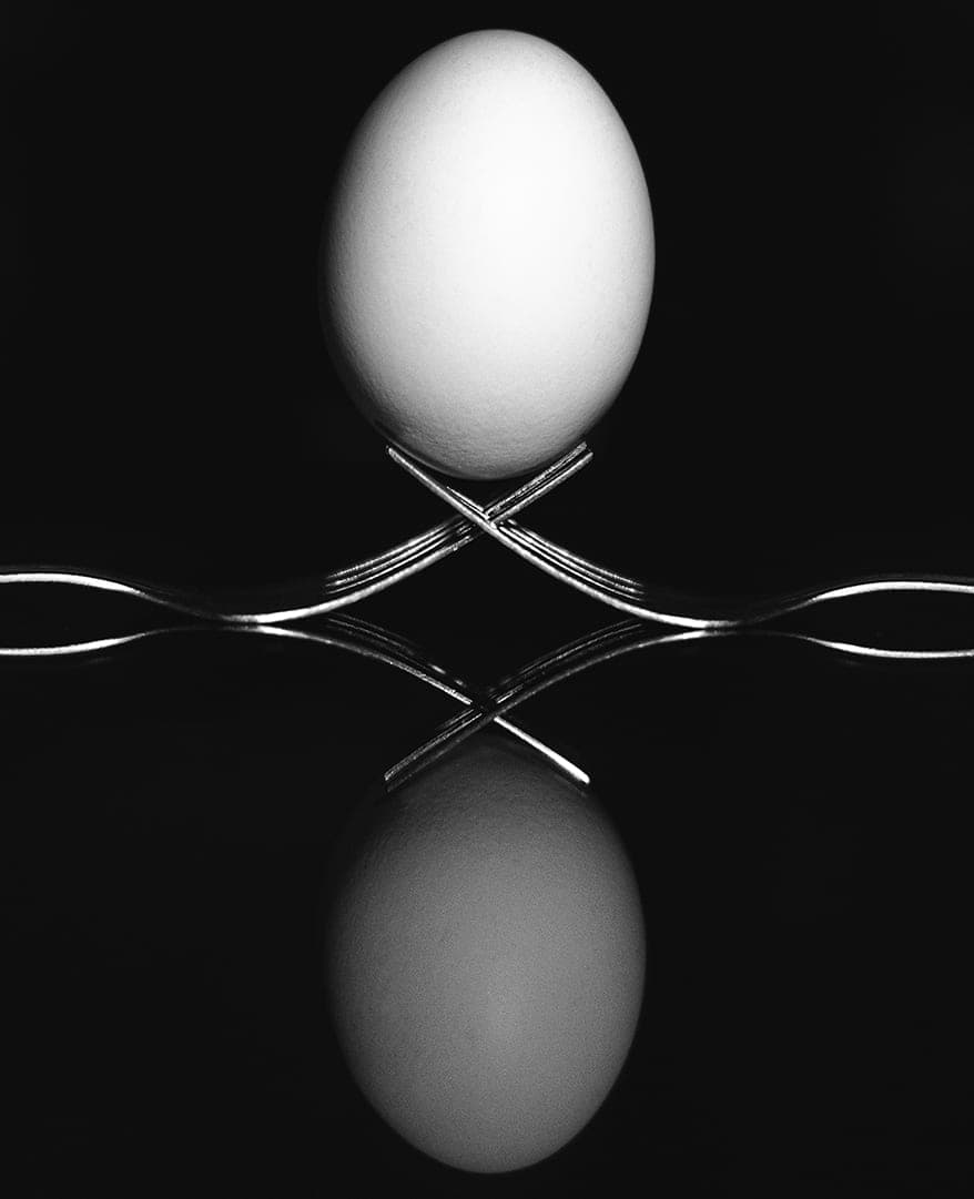 An egg balanced on two crossed forks, mirrored in a dark reflective surface